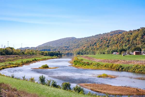 River winding through a grassy valley with tree-covered mountains in the background under a clear blue sky.