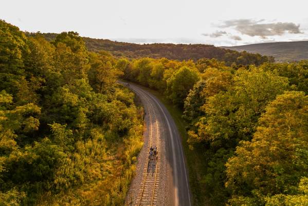 Four passengers peddle along a rail line on peddle bikes with the trees, sky and clouds in the background at sunset.