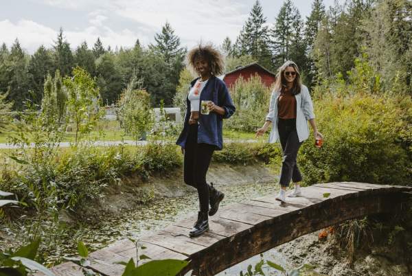 Two women walk across a small bridge, carrying glasses of cider.