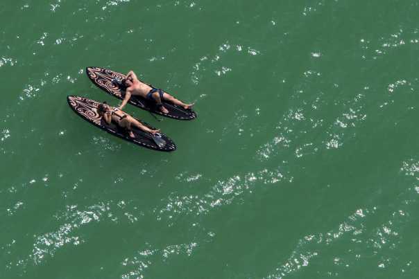 Seen from the air, a man and woman, seemingly in the middle of a green ocean, lie on paddle boards.