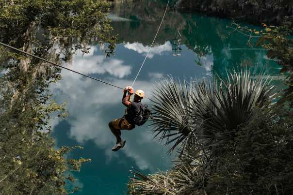 A man connected to a long cable zips above treetops and blue water perhaps 100 feet below.