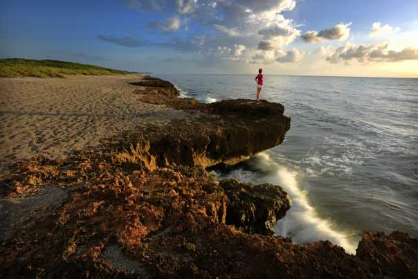 A woman stands on the brink of a brown, craggy rock edge, perhaps 3 feet above the incoming Atlantic Ocean. The surf is reflect the morning light just after dawn.