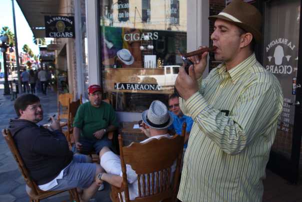 A man in a summer fedora lights a cigar on a city street