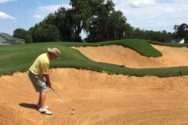 A golfer wearing khaki shorts, yellow polo, and Panama hat prepares to hit a long shot from deep greenside bunker.