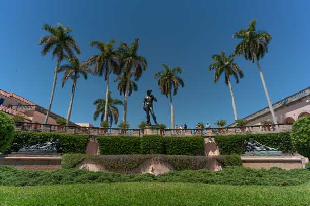A reproduction of Michelangelo's David looms over a vast courtyard of green flora and slender palm trees and Renaissance-style architecture.