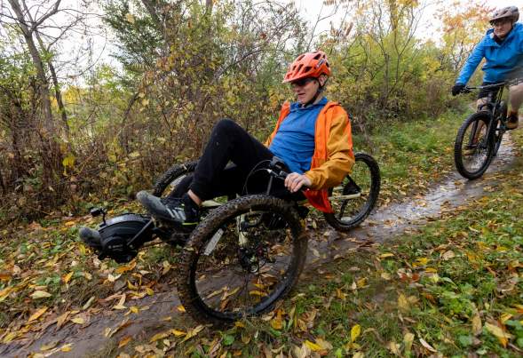 A white man in a low three-wheeled bicycle used to help people with mobility issues rides along a wooded trail