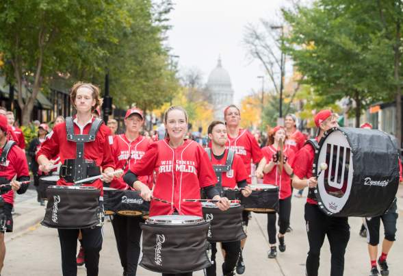 Members of the University of Wisconsin marching band, led by the drum line, march on downtown street with the Capitol building in the background.