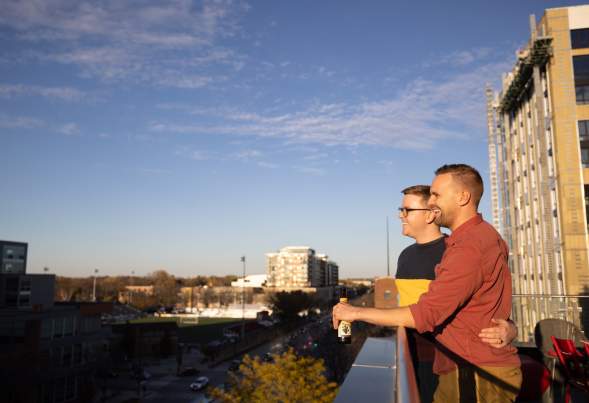 Two men hold hands and look out onto the city from a balcony at the Moxy Hotel