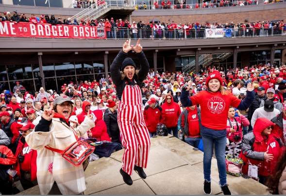Three women dressed in UW badger gear jumping and cheering with a crowd of Badger fans behind them.
