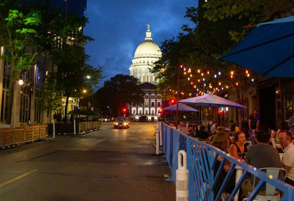 A lively street scene at night, featuring outdoor dining under string lights, with a prominent dome building in the background.