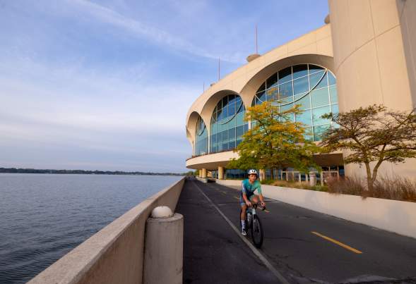 A man in a biker suit wearing a helmet as he rides his bike past the lake and Monona Terrace Convention Center.