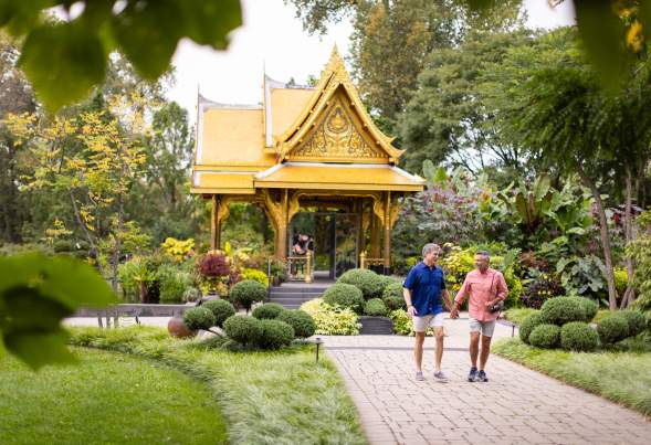 Two men holding hands walk away from the gold Thai pavilion building at Olbirch Botanical Gardens. The couple is smiling at each other and is surrounded by lush greenery.