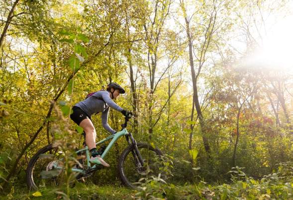 A white woman rides a bike through a wooded trail on a sunny day.