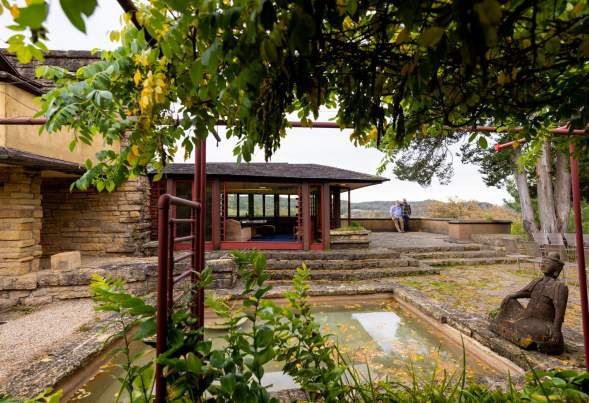 The outdoor courtyard at Frank Lloyd Wright's Taliesin with a reflection pool, Buddhist statue and trees surrounding a room of the home that has large glass windows.