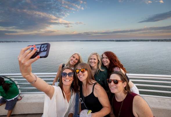A group of six adult women pose for a selfie on a rooftop with Lake Monona in the background