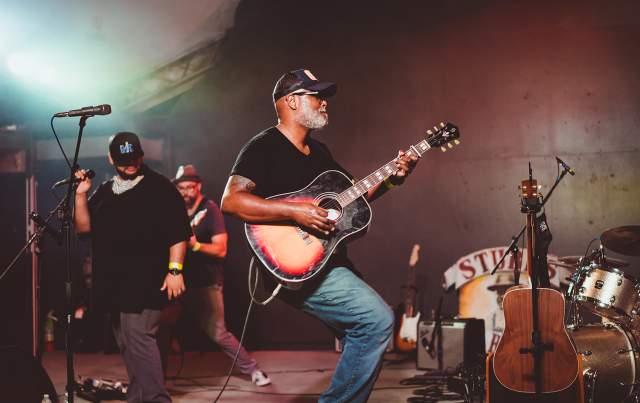 Musician Ray Prim plays an acoustic sunburst guitar on stage at Stubb's in Austin Texas. Two other male musicians stand behind him