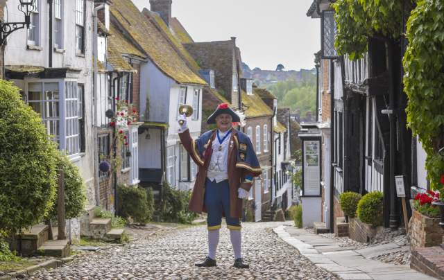 Rye cobbled street with Town Crier holding up his bell wearing traditional costume