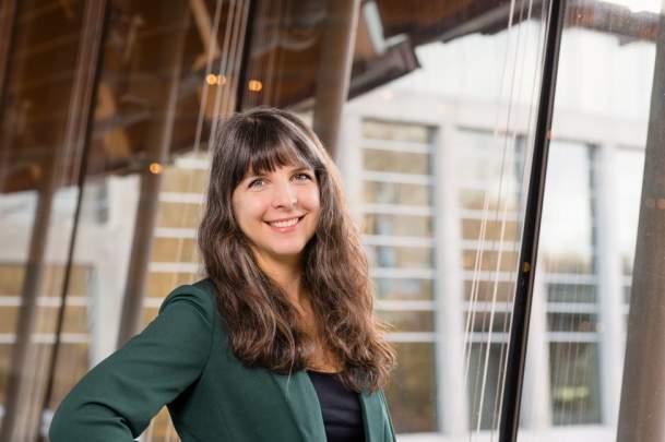 A woman with long brown hair and dressed in a green blazer smiles while standing indoors near a large window.