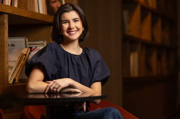 Natalie Stika, wearing a navy blouse, sits at a round table in a cozy setting with wooden bookshelves filled with records and books in the background, smiling warmly at the camera, at the Motto hotel.