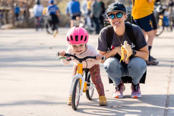 A child wearing a pink helmet learns to ride a balance bike with encouragement from an adult during the Collegiate MTB Championships, surrounded by cyclists.
