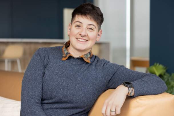 Mer Wilder with short dark hair and a septum piercing, wearing a blue sweater over a plaid collared shirt, sits on a tan couch smiling in a modern indoor space with dark blue walls and greenery in the background.