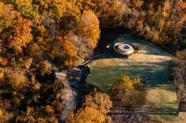 Aerial view of James Turell SkySpace: The Way of Color surrounded by autumn foliage, with winding paths and grassy areas nearby.