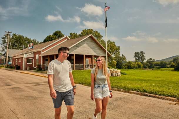 Two people, in summer, walking down a street with a VFW building behind them, and rolling hills.