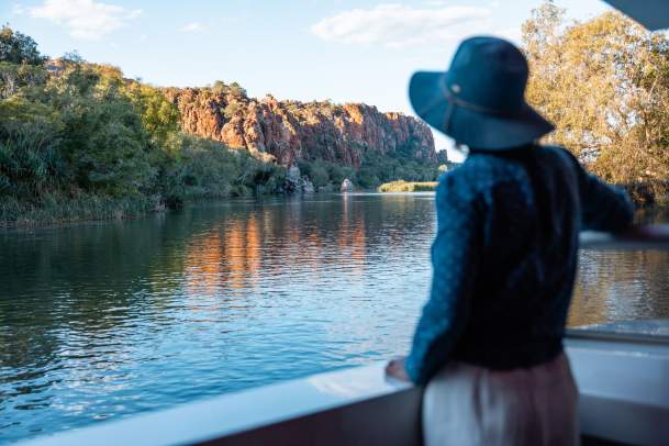 A woman enjoying the scenery on an Ord River cruise - the woman is standing on the vessel looking across the water at a cliff faced bathed in sunlight.