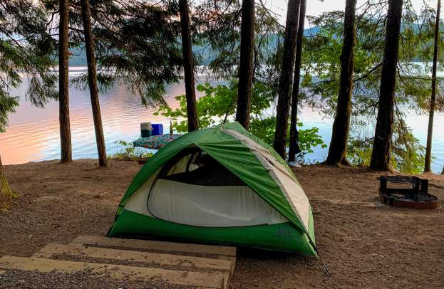 A green and white tent sits beside a lake with tall trees and sunrise colors reflecting off the water.