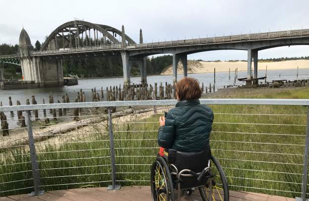 A person in a dark puffy coat with a wheelchair is on the flat boarded viewing point looking out toward the historic Siuslaw River Bridge. The viewing platform has a thin rail and wire fence allowing optimal viewing with the top rail below the person's sightline. Green rushes and grass fill in the space between the deck and riverside. Sand dunes are in the background.