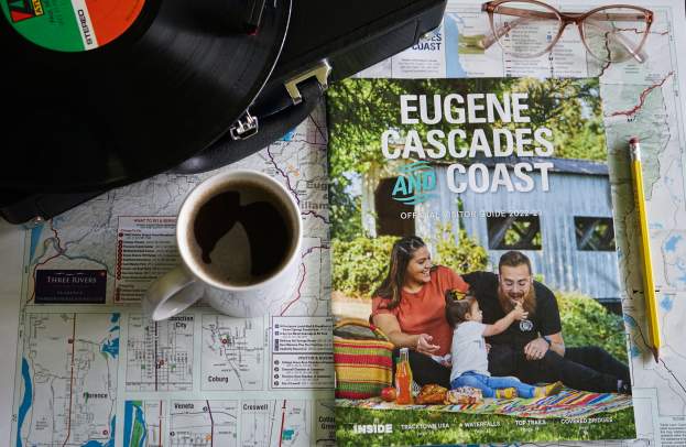 A visitor guide showing a family picnicking near a covered bridge sits on a table with a map, cup of coffee, glasses, a pencil and a record player.