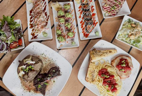 A table full of food plates, including sushi, steak, spaghetti, and more.