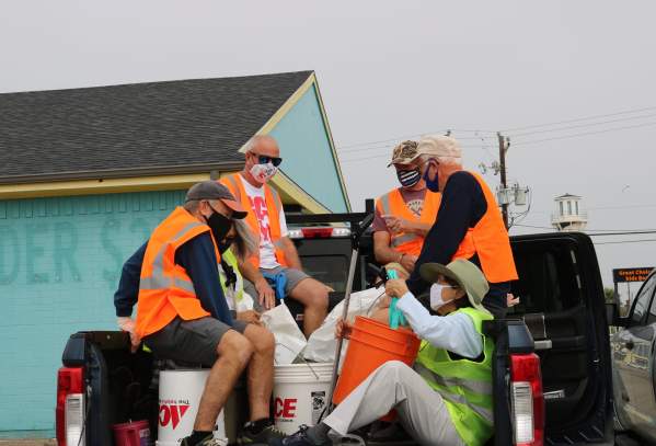 A group of five volunteers in orange safety vests sit in the bed of a truck with trash bags and buckets