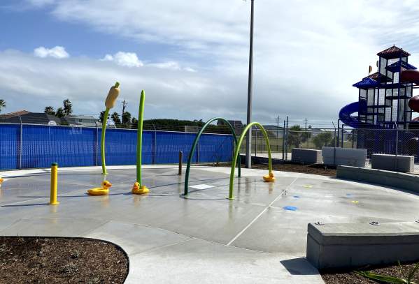 On a gray pad sits several green and yellow hoops and poles with water for kids to splash on. A playground is in the back right.