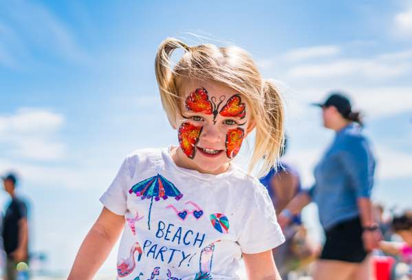 A little girl with blond pigtails and a butterfly-painted face smiles into the camera