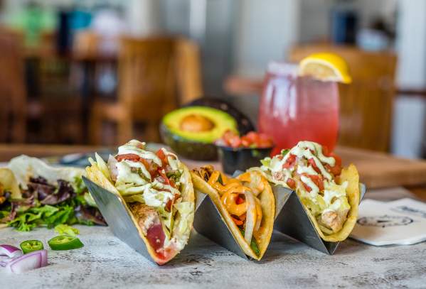 A trio of tacos sits on a table surrounded by an avocado, a pink margarita, and other ingredients