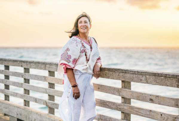 Woman in white and pink linen outfit leans against pier at sunrise