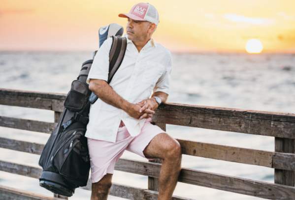 Man carrying golf clubs stands on a pier at sunrise. He's wearing a baseball cap, pink shorts, an white button up, and sneakers.