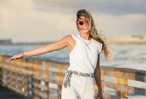 Model wearing an all white outfit with red sunglasses smiles and struts across a pier