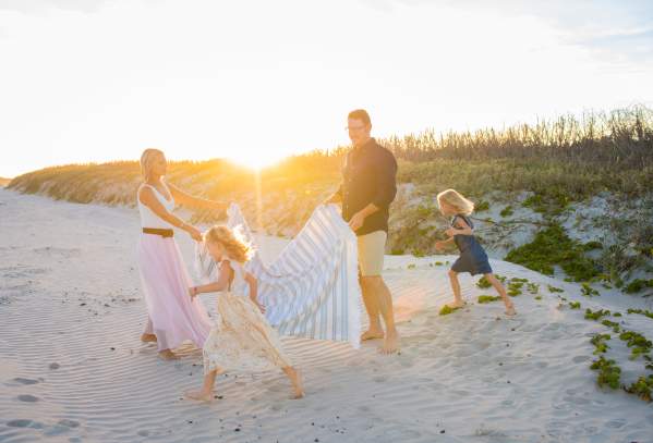 On the beach in front of sand dunes, a mother and father hold a striped blanket between them while two young girls run next to them