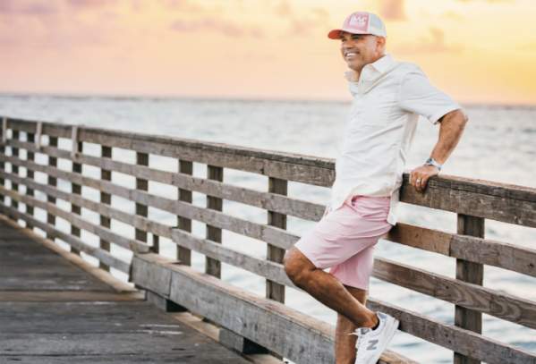 Man in a casual pink and white shorts and shirt outfit leans against a pier at sunrise