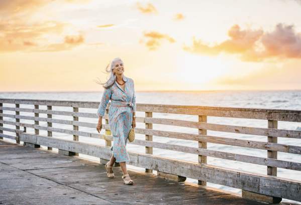 Model walks along a pier at sunrise wearing a turquoise and coral dress and holding a fish purse