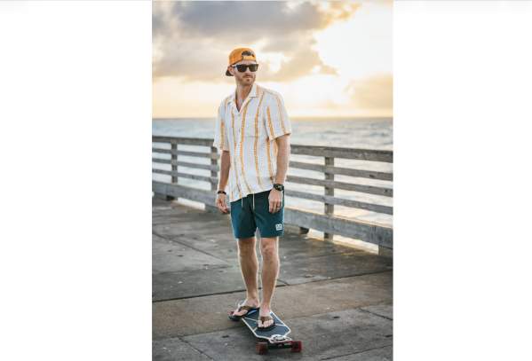 Model in an orange and white striped button up, teal shorts, and a hat rides a skateboard on a pier