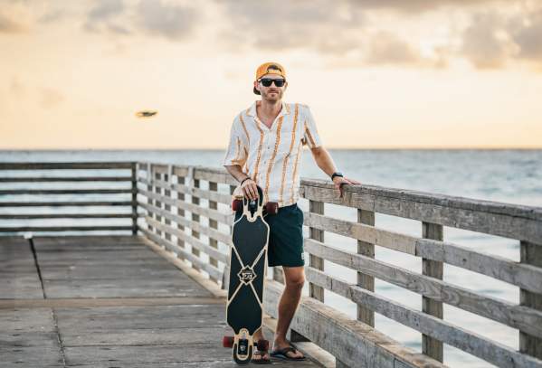 Male model in a casual outfit leans against pier fence while holding on to a skateboard
