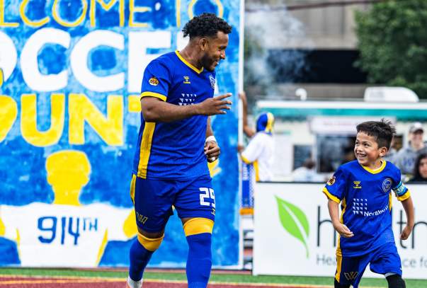 A Westchester Soccer Club player runs out onto the field accompanied by a youth player.