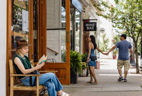 A woman sits on a bench reading a book while a couple in the background is walking into a shop.