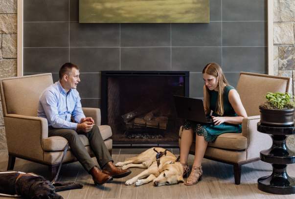 Two people are seated in front of a fireplace with their service dogs lying asleep at their feet. On person has a laptop open and is describing her report to the listener.