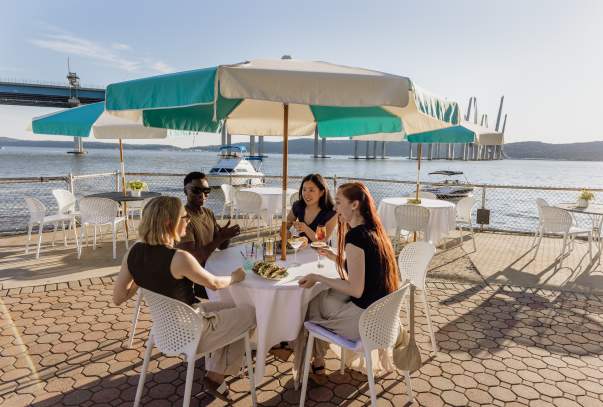 A group of four people sit at an outdoor dining table overlooking the Hudson River. They are laughing and chatting.