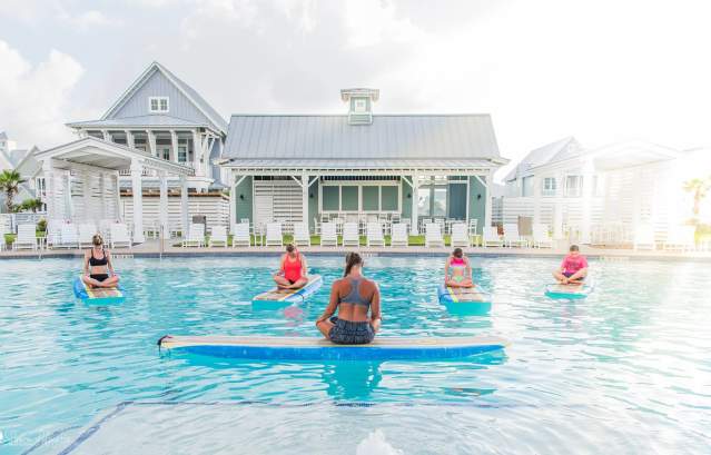 A woman with crossed legs sits atop a surf board floating in a pool, back to the camera. Four women, also with crossed legs, sit atop surf boards facing the instructor.
