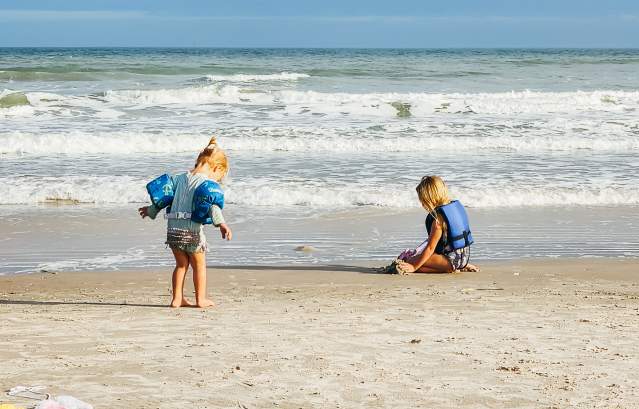 Two blonde toddlers, one in water wings and one in a life jacket, play on the beach by the water. one is kneeling in the sand, one is standing, walking towards the water